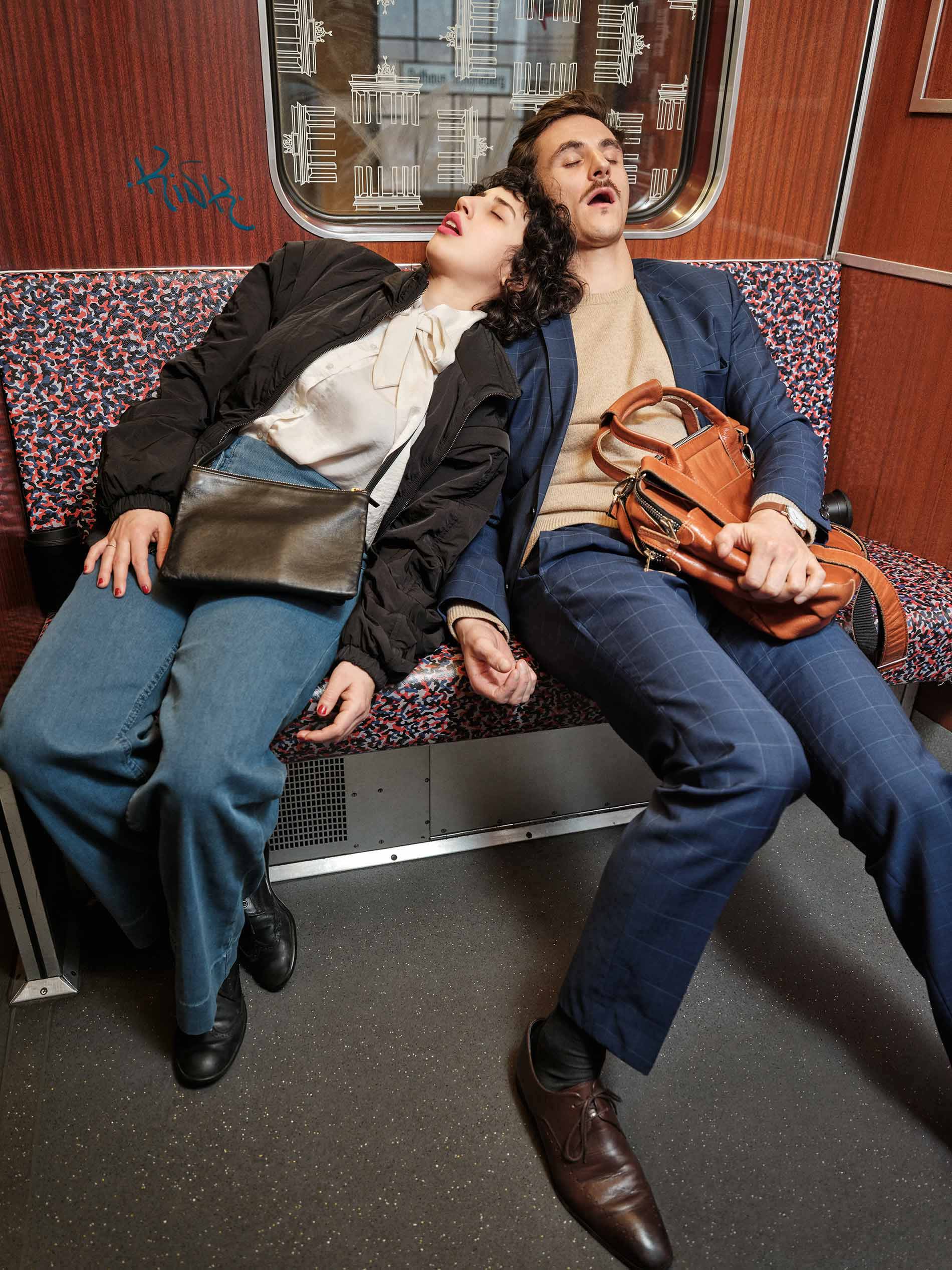A young man and young woman sleep leaning against each other with open mouths and messenger bags in the Berlin U-Bahn | mey®