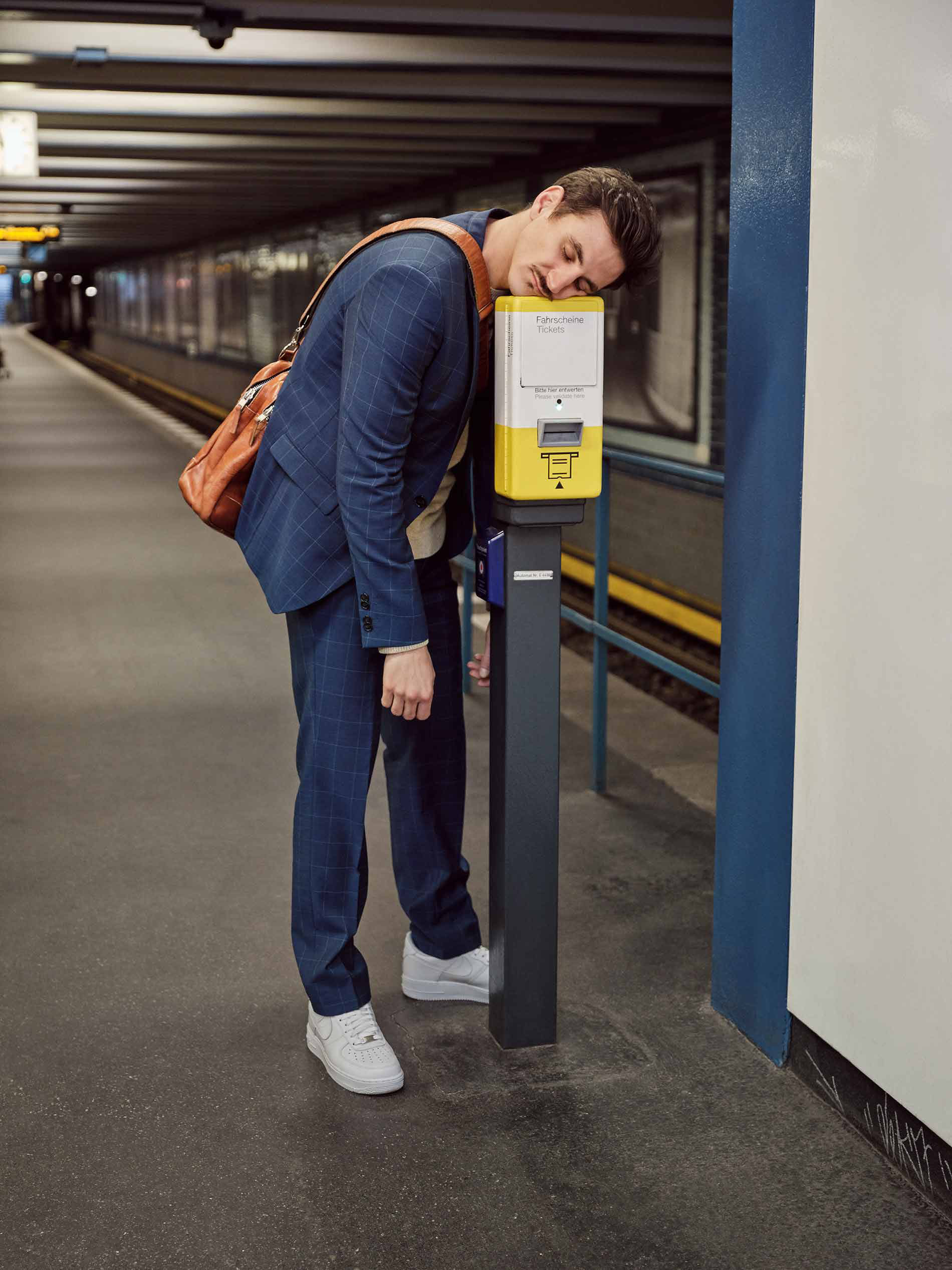 Junger Mann schläft im stehen an einem U-Bahnhof in Berlin mit dem Kopf auf den Fahrkarten-Entwertungsautomat gelehnt | mey®
