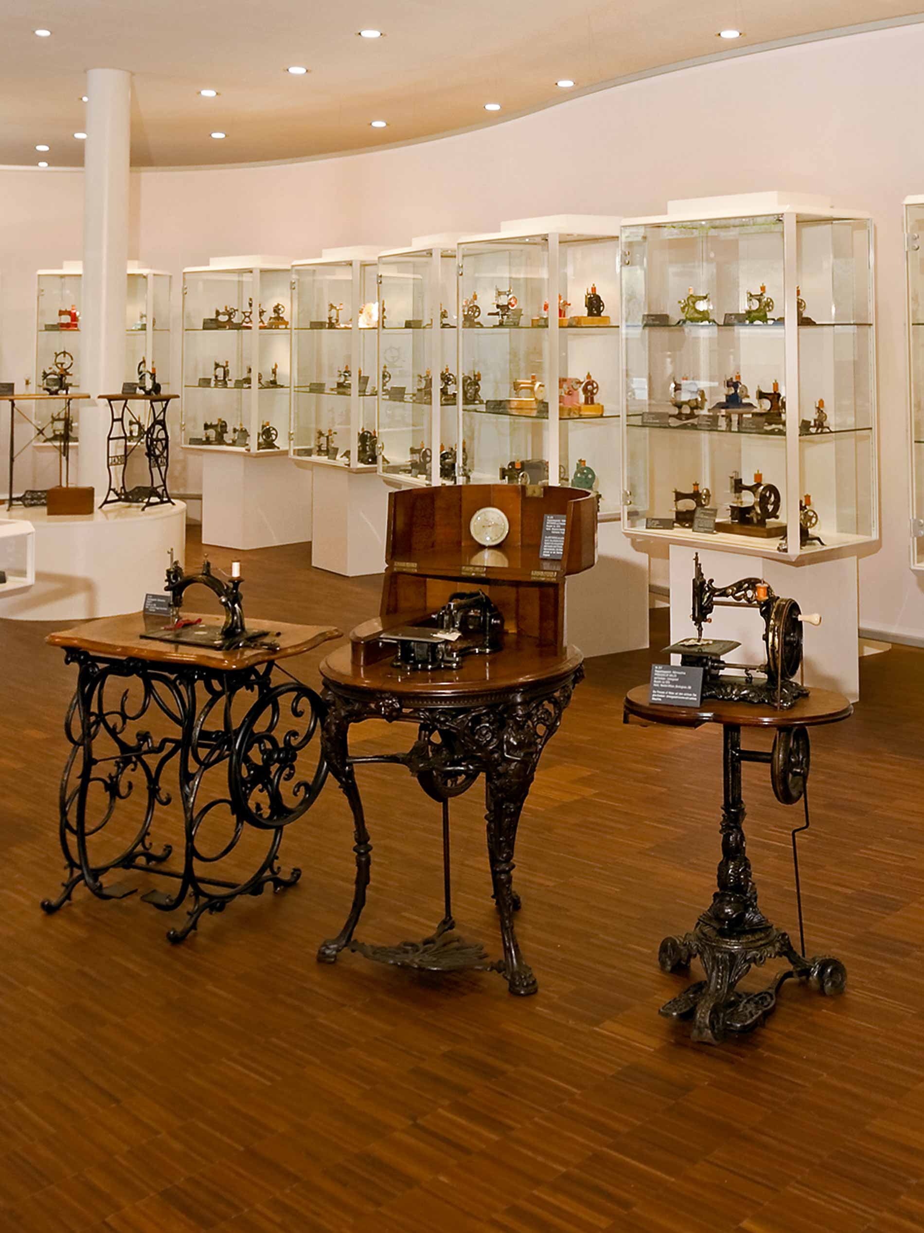 Innenraum des Museums, drei historische Nähmaschinen im Vordergrund und mehrere Glasvitrinen mit Ausstellungsstücken im Hintergrund