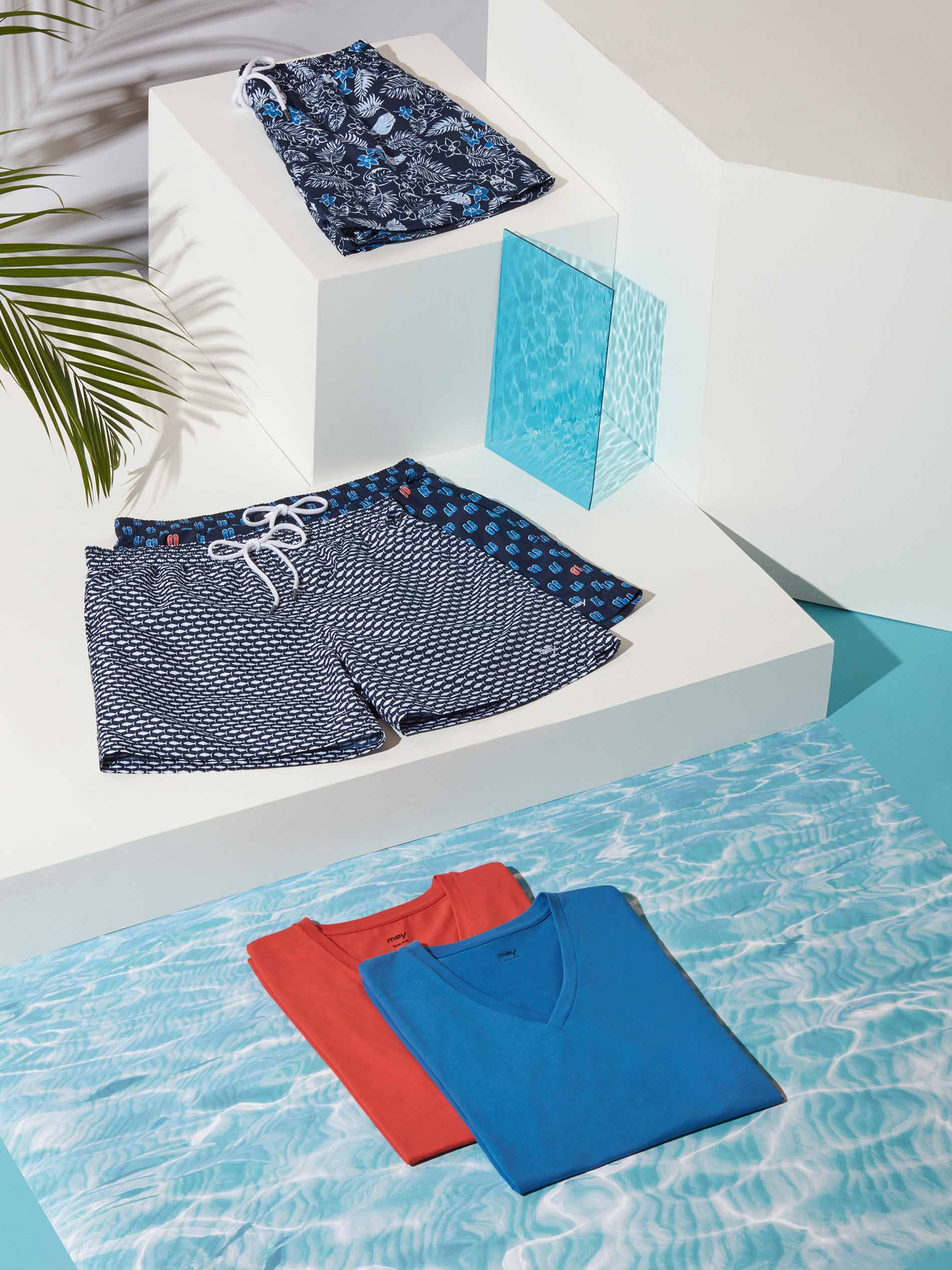 Swimwear Badeshorts und farbige T-Shirts arrangiert auf weißen Stelen mit Wasser-Hintergrund und Palmenblatt | mey® 